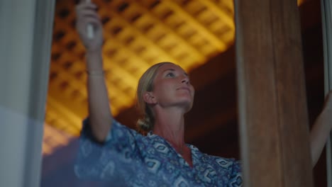 mujer en albornoz de seda azul cerrando las cortinas de la habitación del hotel, lista para la noche