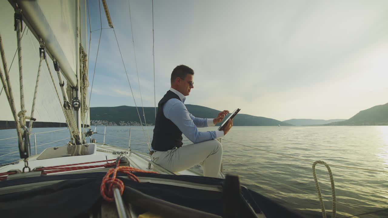 Uomo che legge un tablet su una barca a vela al tramonto