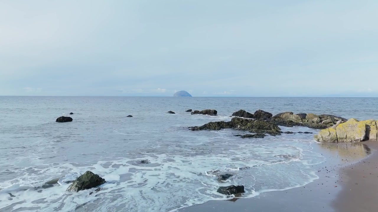 Wellen brechen über Felsen an der Küste von Ayrshire, mit Ailsa Craig am Horizont sichtbar.