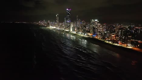 Luces-De-La-Ciudad,-Paraíso-De-Los-Surfistas-A-Cabezas-De-Burleigh,-Volando-Alto-Sobre-El-Mar-Mirando-Hacia-Atrás-Al-Paraíso-De-Los-Surfistas,-Paisaje-Marino-Nocturno,-Olas-Brillantes