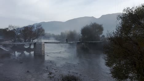 tourist crossing old rope bridge over misty chinese river, slider aerial view