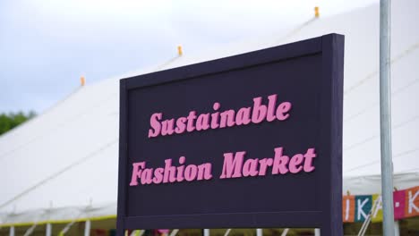 pink text sign written sustainable fashion market on a purple board marking a place for recycled clothing made by trusted environmental activists placed in the front of a massive tent festival