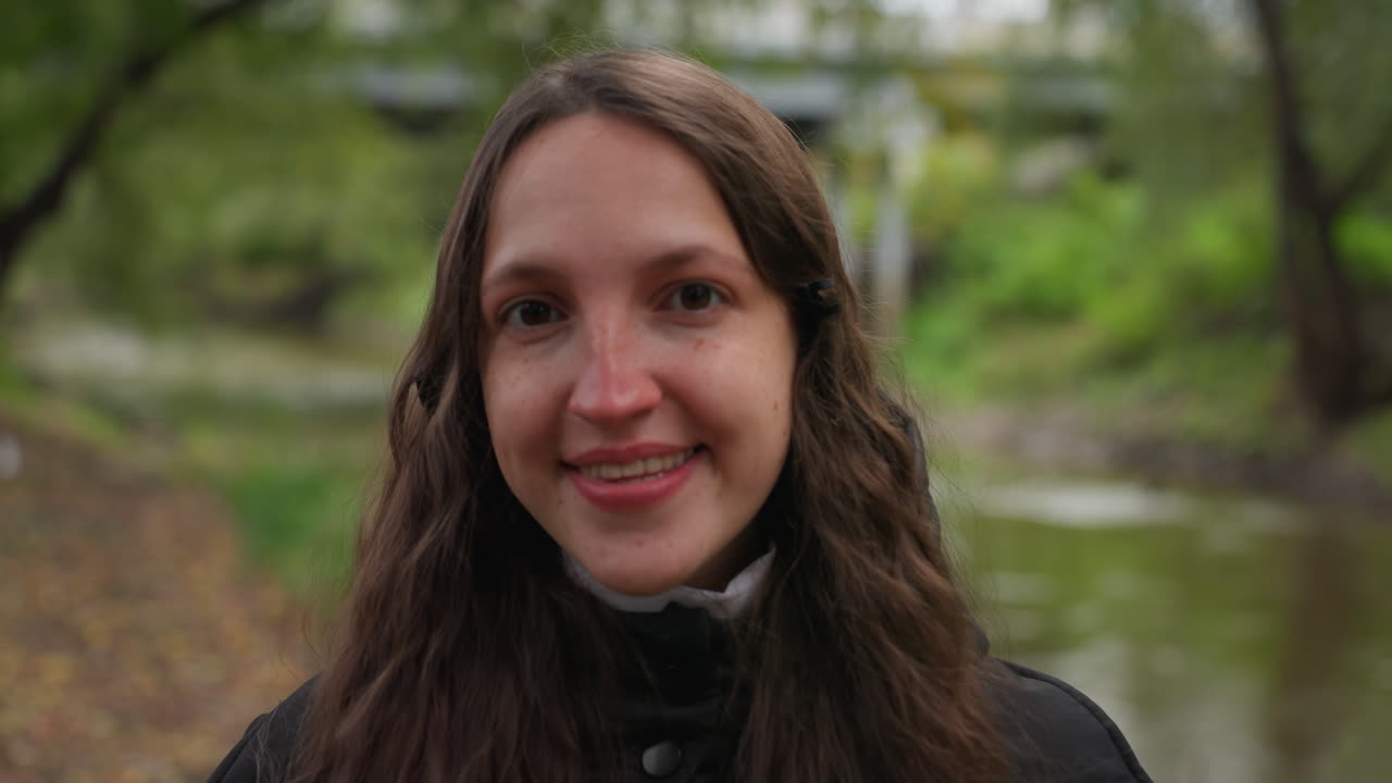 Ritratto ravvicinato di donna caucasica sorridente in riva al fiume, ponte sullo sfondo, bokeh delicato, lunghi capelli castani, cappotto nero, foglie autunnali, espressione calorosa, sguardo amichevole verso la macchina fotografica, luce naturale, foto per documenti da esterni
