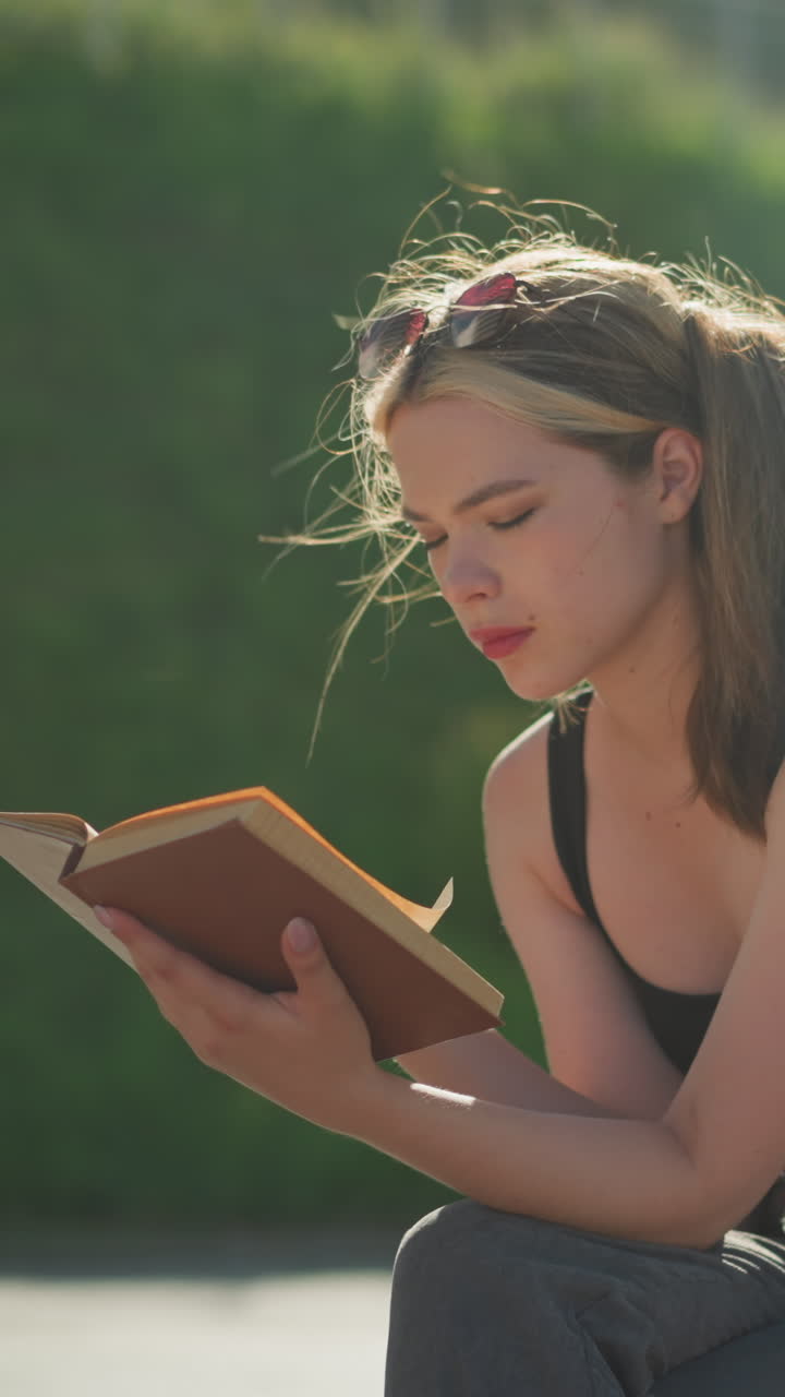 dama sentada al aire libre lee un libro mientras el viento sopla a través de su cabello y páginas del libro, con la luz del sol reflejándose en ella, el fondo borroso incluye una escalera y un hombre que pasa en una tapa azul