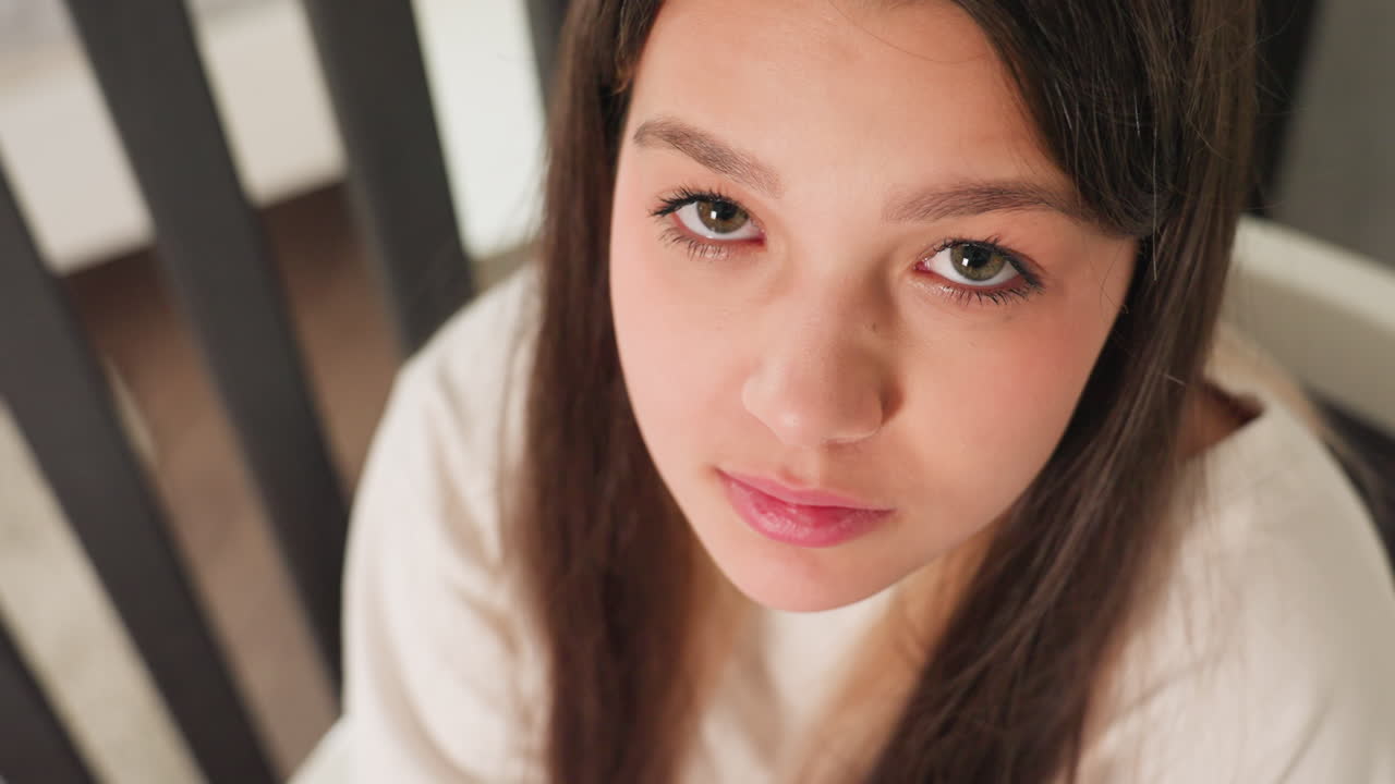 Aerial view beautician seated indoors, eyes down then lifting gaze, long brunette hair, soft light on natural skin, cozy room background with bed and rails, contemplative portrait moment