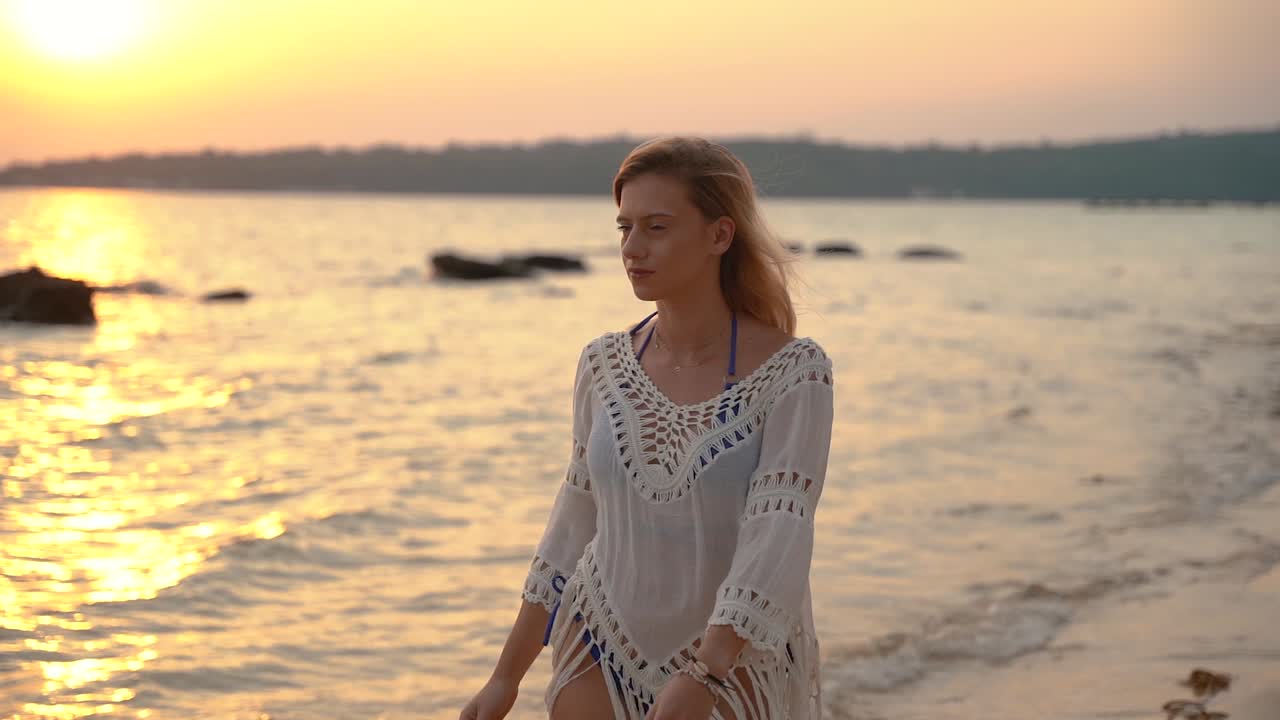 hermosa joven europea en bikini azul y camisa blanca camina en el surf durante el amanecer en una playa de arena en tailandia mientras mira alrededor