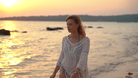 beautiful young european woman in blue bikini and white shirt walks in the surf during sunrise on a sandy beach in thailand while looking around