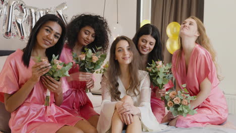 group of multiethnic female friends and bride who is wearing white silk nightdresses looking at camera sitting on bed holding bouquets decorated with word 'bride' ballons in bridal gathering