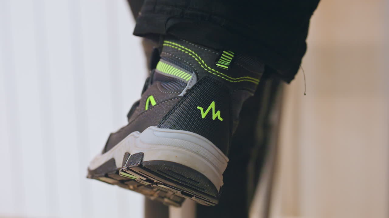 Primo piano di una sneaker da bambino che dondola sul grembo della madre, scarpa con dettagli neon e suola testurizzata. Il piedino del bambino pende delicatamente dalla sedia, in una luce interna accogliente. Un momento genitoriale giocoso con una sottile sfocatura di movimento e un'atmosfera calda.