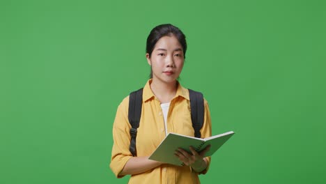 asian woman student with a backpack reading a book then smiling to camera while standing in the green screen background studio