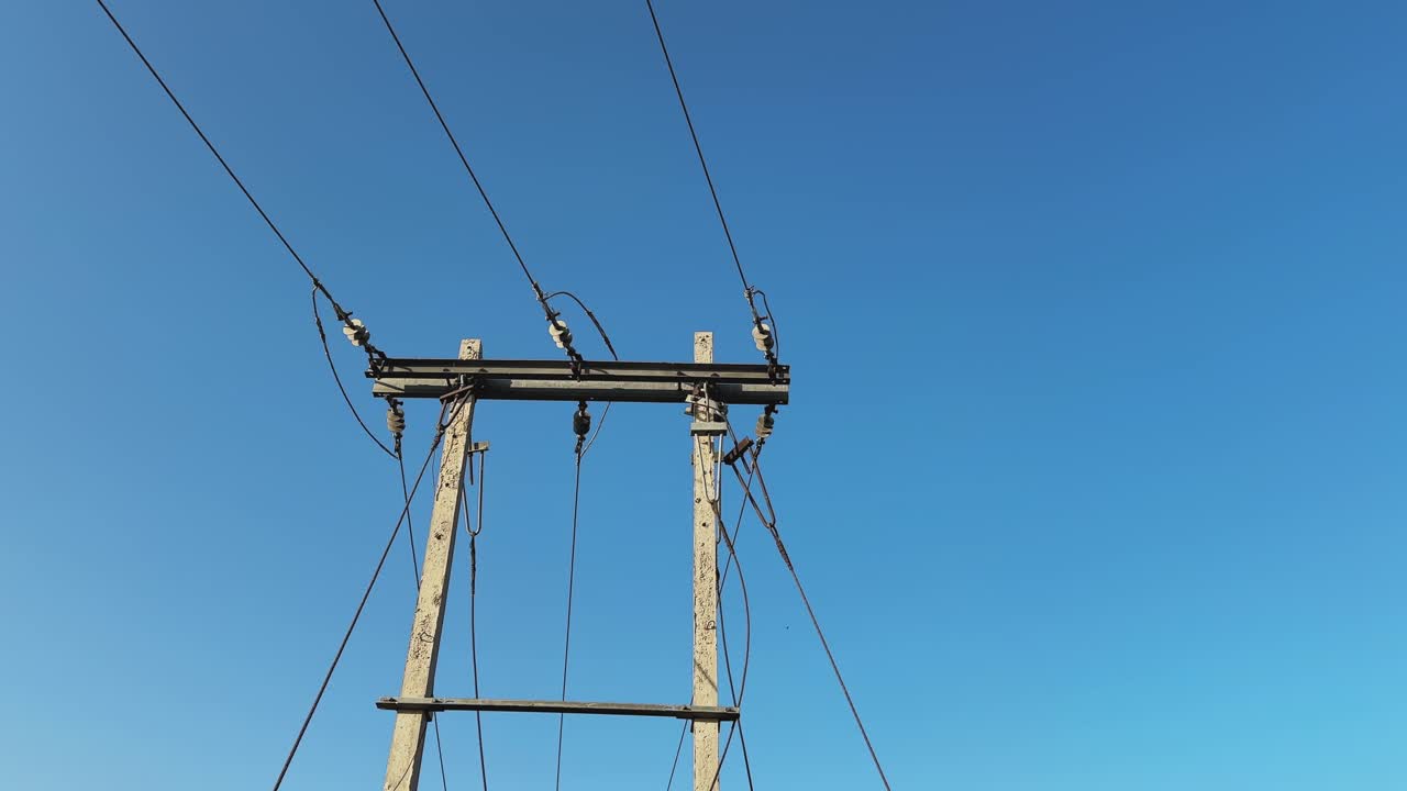 Slow orbiting shot around a PSC or reinforced cement concrete power distribution pole in India, overhead electrical lines and steel crossarm silhouetted against a clear blue sky