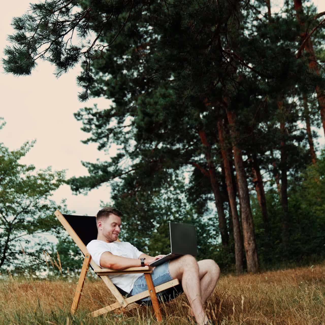 Uomo caucasico felice che lavora da remoto. Imprenditore in T-shirt e jeans seduto su una sedia pieghevole con un portatile in mano. Natura sullo sfondo. Vista dal basso