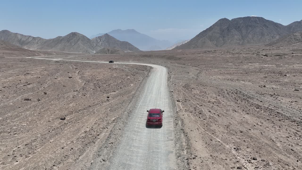 toma aérea de un vehículo rojo y negro conduciendo por una carretera polvorienta en el desierto de perú frente a la ciudad santa de caral con colinas, montañas y rocas en un día soleado