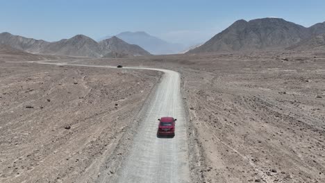 aerial dolly shot of a red and black vehicle driving along a dusty highway in the desert of peru in front of the holy city of caral with hills, mountains and rocks on a sunny day