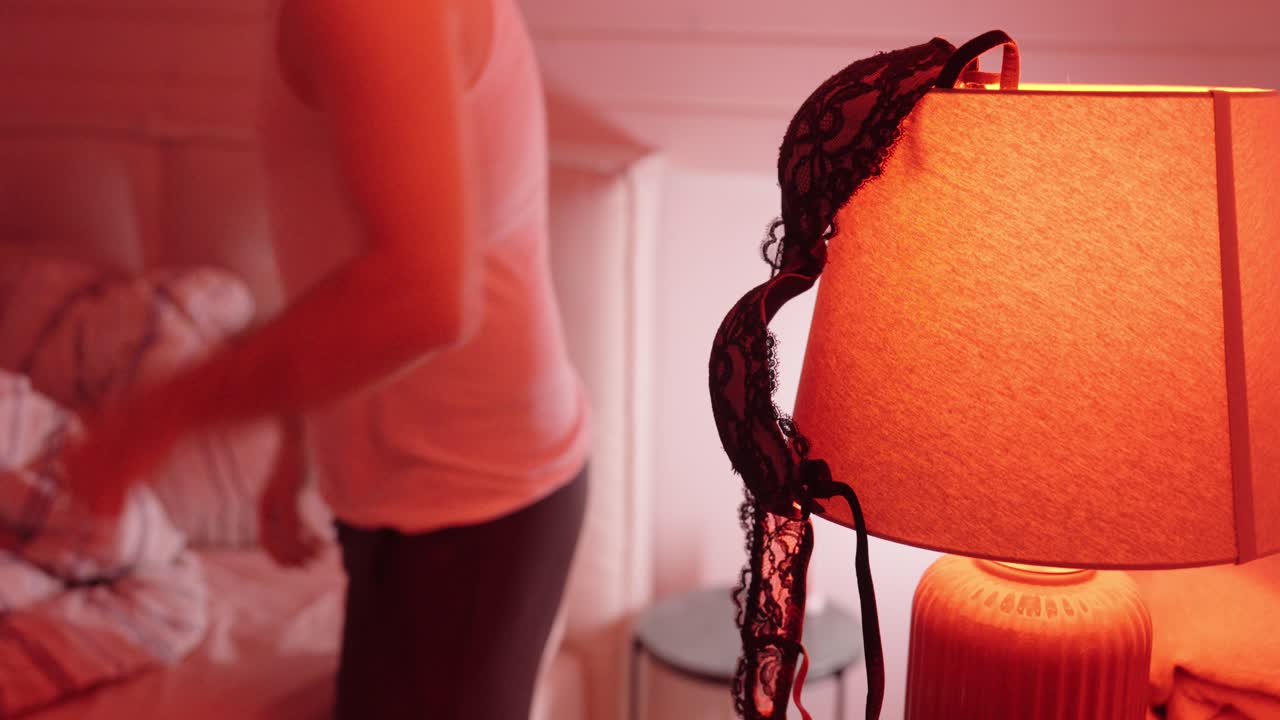 A dimly lit bedroom scene with lingerie draped over a bedside lamp while a person moves in the background. Concept of intimacy, temptation, secrecy, and relationship tension