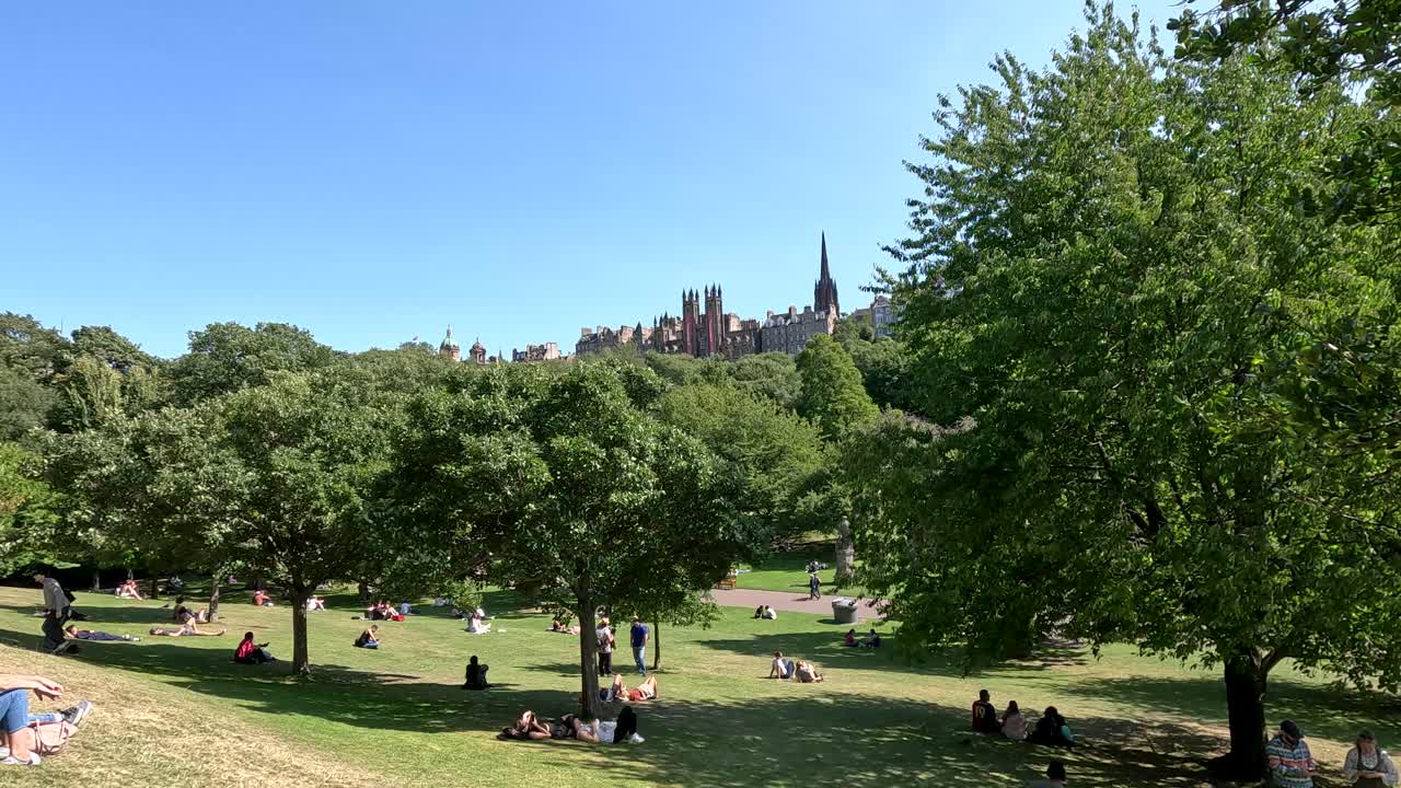Groups of people sit and lounge on grass in a sunlit Edinburgh park, with lush green trees and historic city buildings visible under clear blue skies. Static wide shot