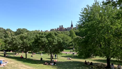 groups of people sit and lounge on grass in a sunlit edinburgh park, with lush green trees and historic city buildings visible under clear blue skies. static wide shot