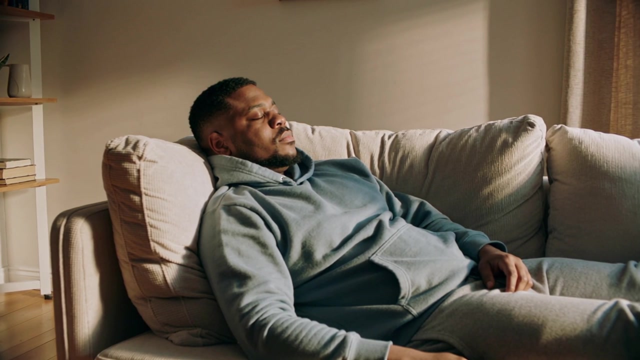 Camera widening, man wearing light-gray hoodie reclining on living-room sofa, resting near shelf