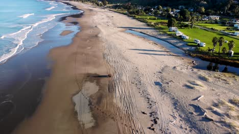 Quad-bike-driving-on-sandy-beach,-aerial-panoramic-reveal-of-New-Zealand-coastal-scenery-of-Tokomaru-Bay