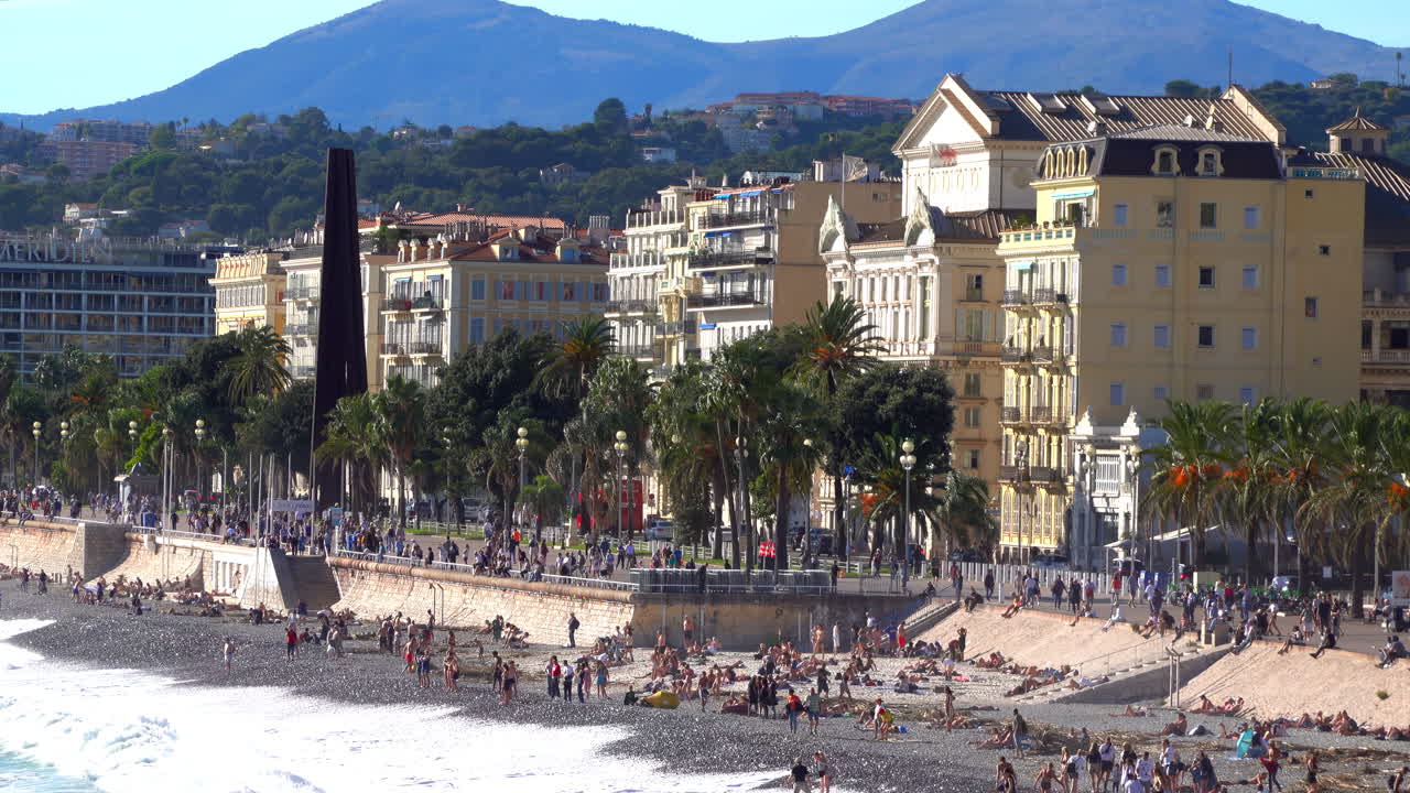 Nice, France - July 3, 2025: Wide view of the Promenade des Anglais with palm trees, historic buildings, and people enjoying the pebble shoreline