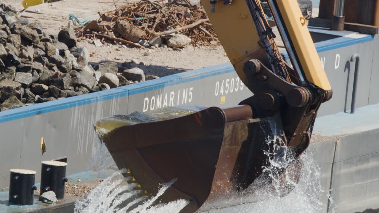 A large excavator lifts and drains water from its bucket beside a river barge during daylight construction in Dresden, Germany. Sunlit, close-up, steady camera