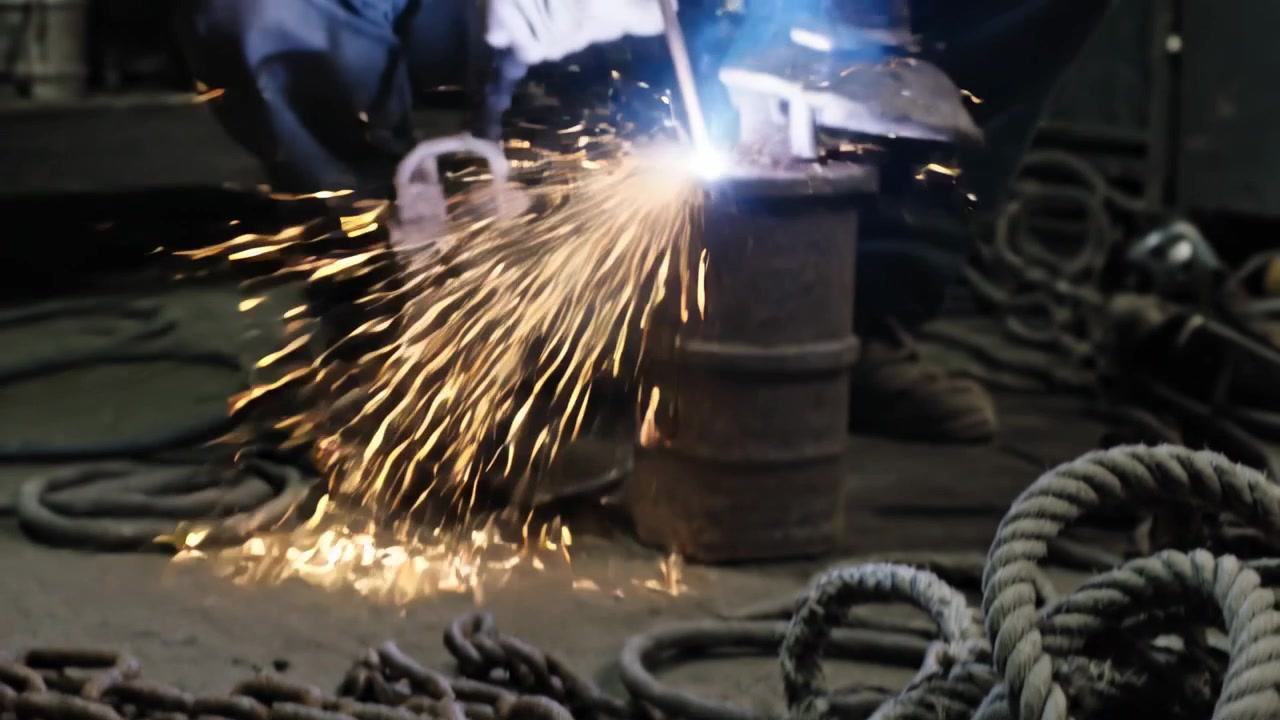 Striking electrode sparking arc, welder crouching and repairing metal in dim shop, leather apron