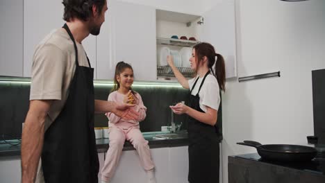 Happy-brunette-girl-in-a-white-T-shirt-and-black-apron-together-with-her-husband-with-brunette-stubble-and-little-daughter-in-pink-clothes-are-preparing-for-breakfast-and-laying-out-plates-and-food-in-the-morning-in-a-modern-apartment