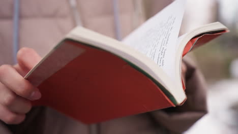 close-up view of person s hands opening red book outdoors, visible printed text on page, soft focus background with hints of winter clothing and natural light, capturing quiet reading moment