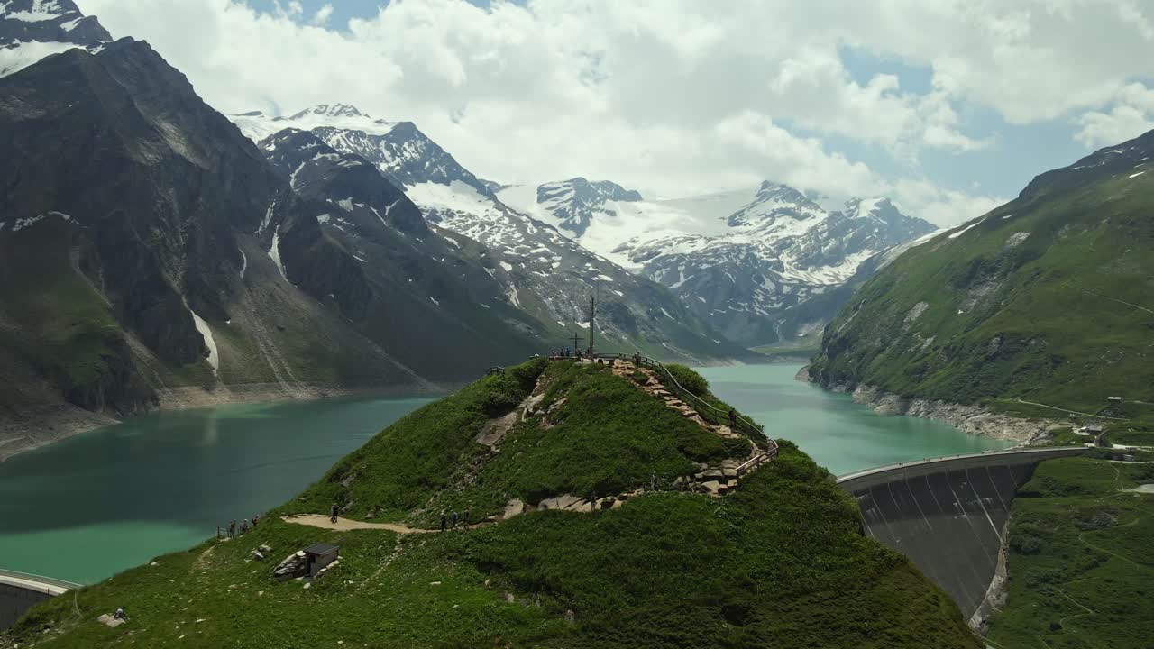 Panoramic view of a turquoise alpine lake surrounded by snow-capped mountains and green valleys. In the foreground, a small hill with a lookout cross overlooks a curved dam embedded in the landscape