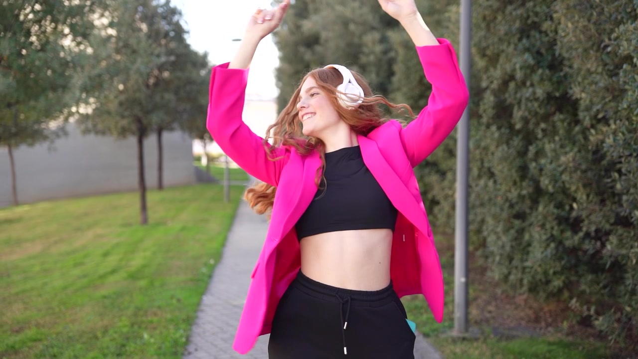 Una mujer disfrutando felizmente de la música al aire libre, mientras lleva una chaqueta rosa vibrante en la naturaleza.