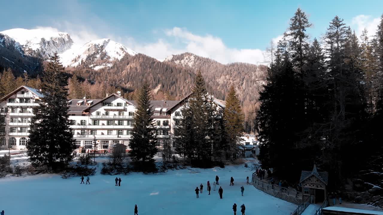 panorámica aérea en cámara lenta de personas caminando en el lago congelado braies cerca de un gran hotel con los dolomitas italianos en el fondo