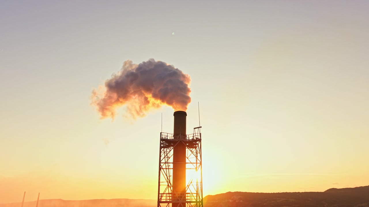 Fumo che sale da un'alta ciminiera industriale contro un cielo sereno durante il tramonto in un'area urbana