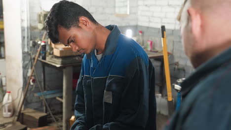 automotive engineers in workshop, one in blue uniform focused on task, colleague showing something, surrounded by mechanical equipment