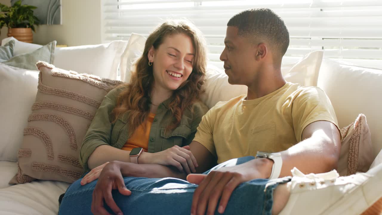 Diverse couple exchanging gentle smiles and calm words, forging cozy bond on living room couch