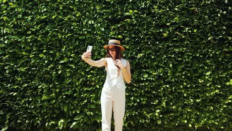 chic woman in white vest and hat takes selfie in vivid green formal garden hedge