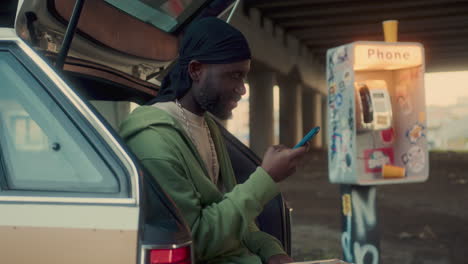 black man using smartphone by car near sticker-covered payphone booth