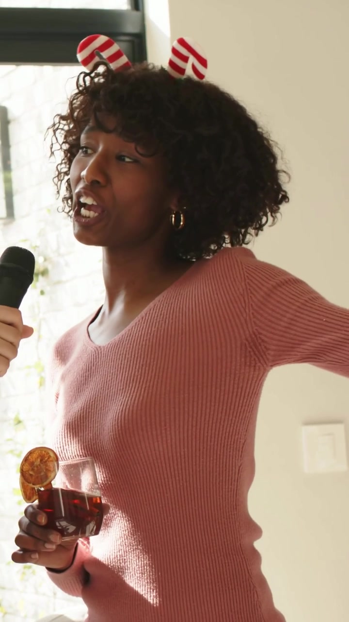 Vertical video: Singing woman in pink top holding wine glass at home, mic entering frame for guests