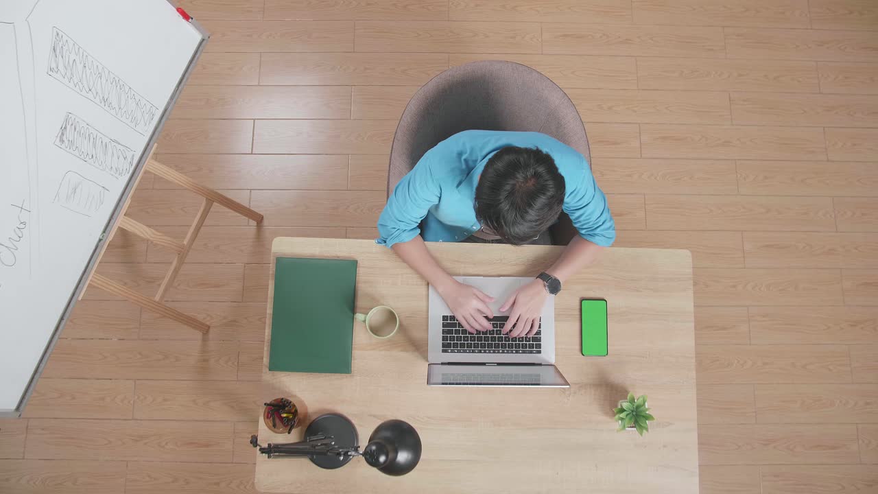 위쪽 뷰  아웃 은 집에서 화이트 보드 에 나무 책상에 앉아있는 동안 녹색 화면 스마트폰 에 노트북에서 타이핑하는 남자의 입니다.