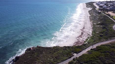 Descending-Aerial-View-Over-Quinns-Beach-With-Waves-Lapping-Shore-And-Walkers-Using-Coastal-Path