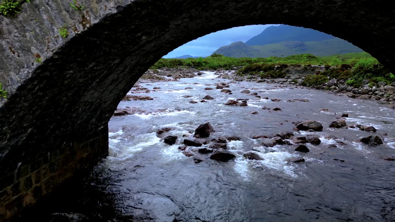 Volo cinematografico con drone sotto lo storico ponte di pietra di Sligachan che rivela la valle delle Highlands scozzesi. Fiume che scorre sotto gli archi con un drammatico paesaggio montano oltre. Ripresa aerea epica sull'Isola di Skye