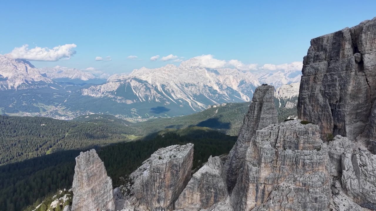 Vista aerea con drone delle formazioni calcaree delle Cinque Torri vicino al Passo Giau, circondate da ghiaioni scoscesi, pendii boscosi e ampie cime dolomitiche sotto un cielo azzurro e limpido.