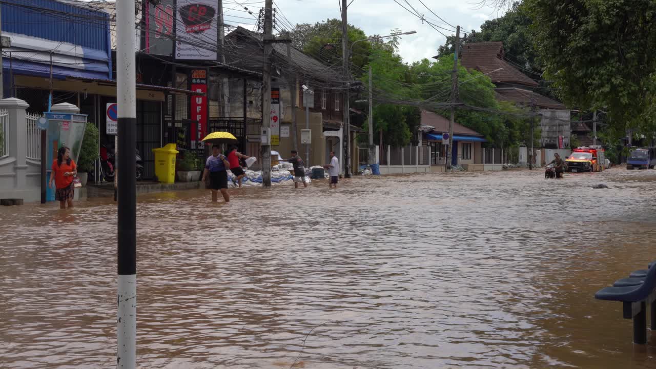 Various individuals are seen helping each other navigate a severely submerged road, where brown water reaches knee height A fire and rescue truck drives by community support flooding situation
