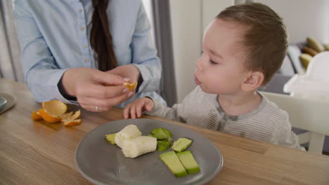 Unerkennbare-Mutter,-Die-Ihren-Kleinen-Sohn-Mit-Clementine-Füttert,-Während-Sie-Zusammen-Am-Tisch-Im-Wohnzimmer-Sitzt