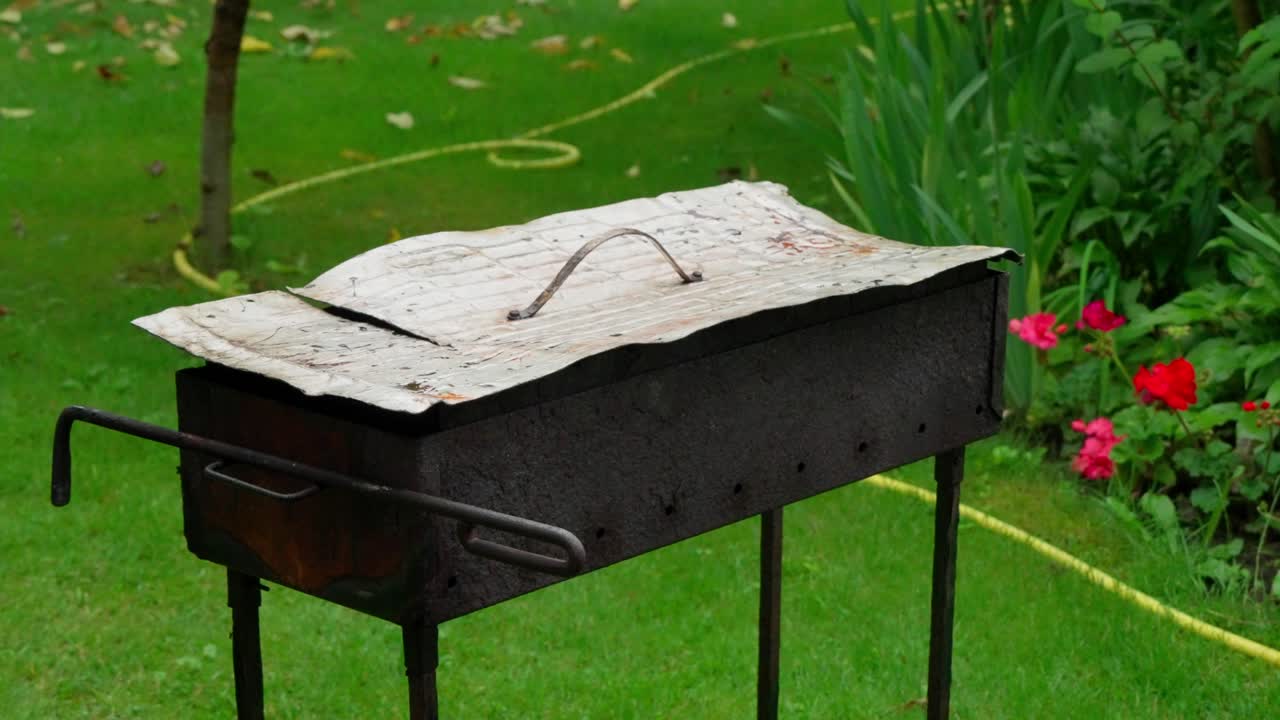 Rusten metallkullgrill står på grønt gress i hagen i Daugavpils. Håndlaget grillutstyr står nær røde blomster og hageslange på en sommerdag for utendørs matlaging