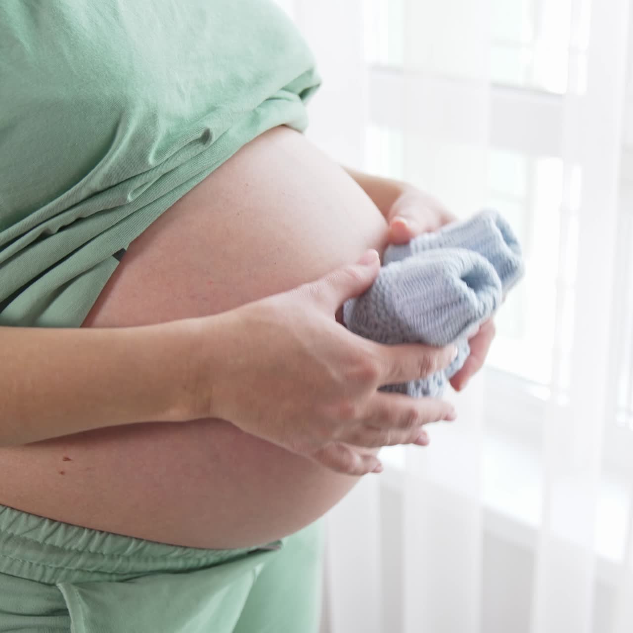 Donna incinta non identificata posiziona piccoli calzini da neonato sulla parte superiore della sua grande pancia e li sposta lentamente verso il basso. La futura mamma gioca con scarpine da neonato lavorate a maglia. Primo piano.