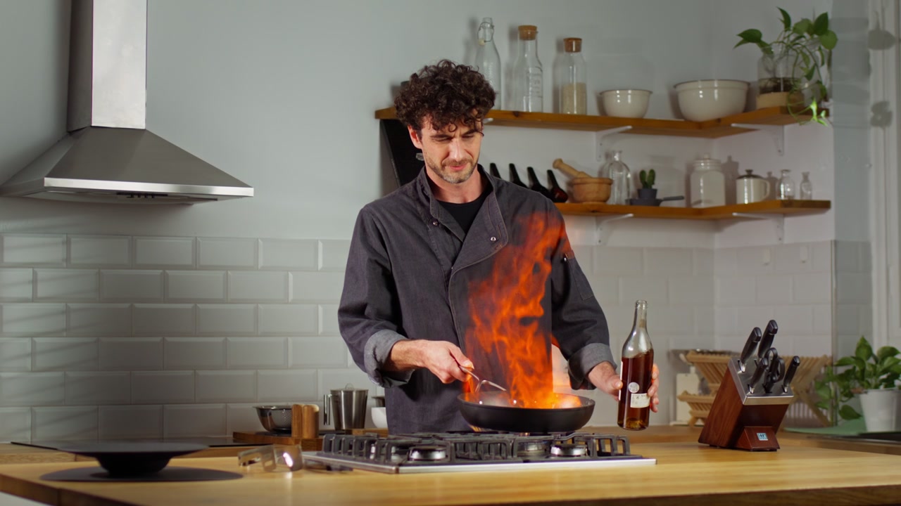 Male cook pours alcohol into a frying pan, creating a dramatic burst of flames during a slow motion flambé in a home kitchen.