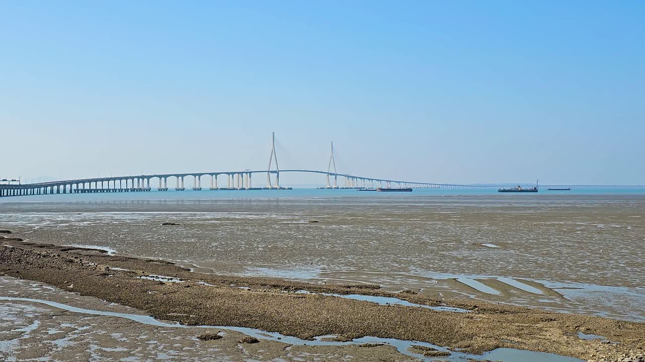 The Incheon Bridge stretches majestically over the sea, connecting the mainland to Yeongjong Island, with its iconic cable-stayed design visible against the clear blue sky, low tide, container ships