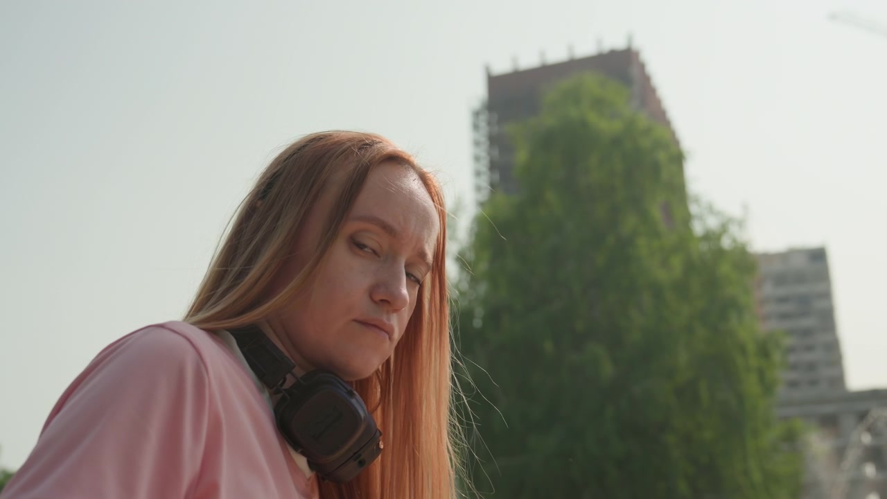 Mulher de cabelo comprido veste camisa rosa e fones de ouvido, em pé ao ar livre em um ambiente urbano. Ao fundo, árvores verdes e edifícios altos sob a luz do dia, criando uma atmosfera calma.