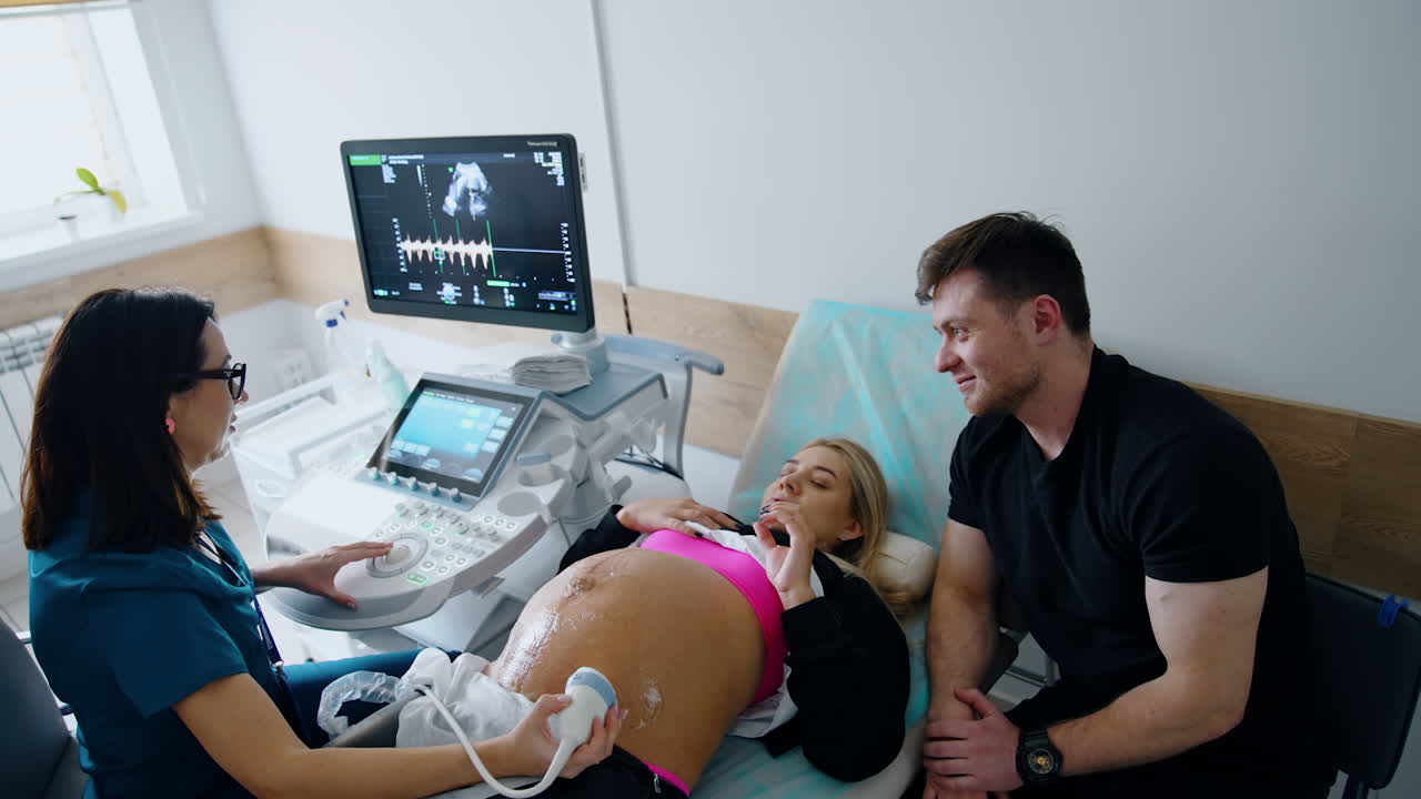 Pregnant woman talks to the doctor during ultrasound examination. Smiling man sits beside listening. Top view.
