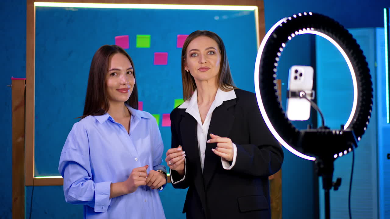 Two Caucasian women stand indoors in front of a camera with light ring. Ladies taking video for a blog. Glass board with sticker notes at backdrop.